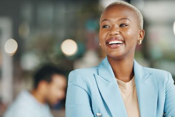 A smiling person in a light blue blazer stands confidently with arms crossed in an office setting, while another individual works in the blurred background.
