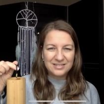 A woman with long brown hair smiles while holding the "Overwhelm to Calm – 1:1 Guided Meditation" product. Behind her, a dreamcatcher with feathers hangs against a dark background.