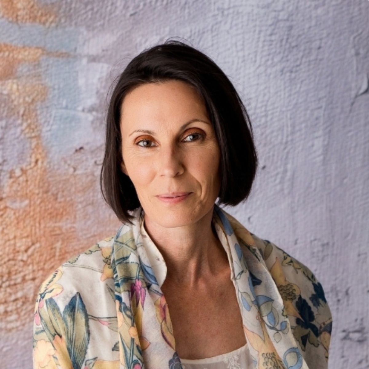 A woman with short dark hair in a light floral shawl stands before a pastel backdrop, calmly gazing at the camera—conveying the tranquil vibe of our Pack of 4 Kinesiology Sessions.