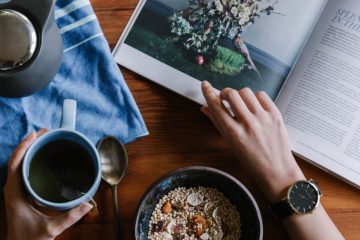 A persons hand pointing at a bowl of cereal and a book.