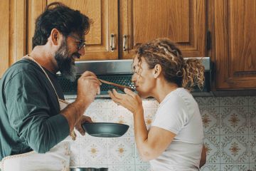 A man and woman stand in a cozy kitchen with wooden cabinets. The man, wearing an apron, offers a spoonful of food to the woman. Both are smiling and look happy. A bowl of fruit and vase of flowers sit on the counter nearby.