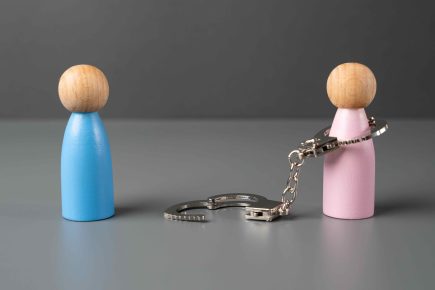 Two wooden figurines, one blue and one pink, stand apart. The pink figurine is attached to an open set of metal handcuffs—symbolizing the journey of healing from a toxic relationship—as the other cuff lies on the ground between them.