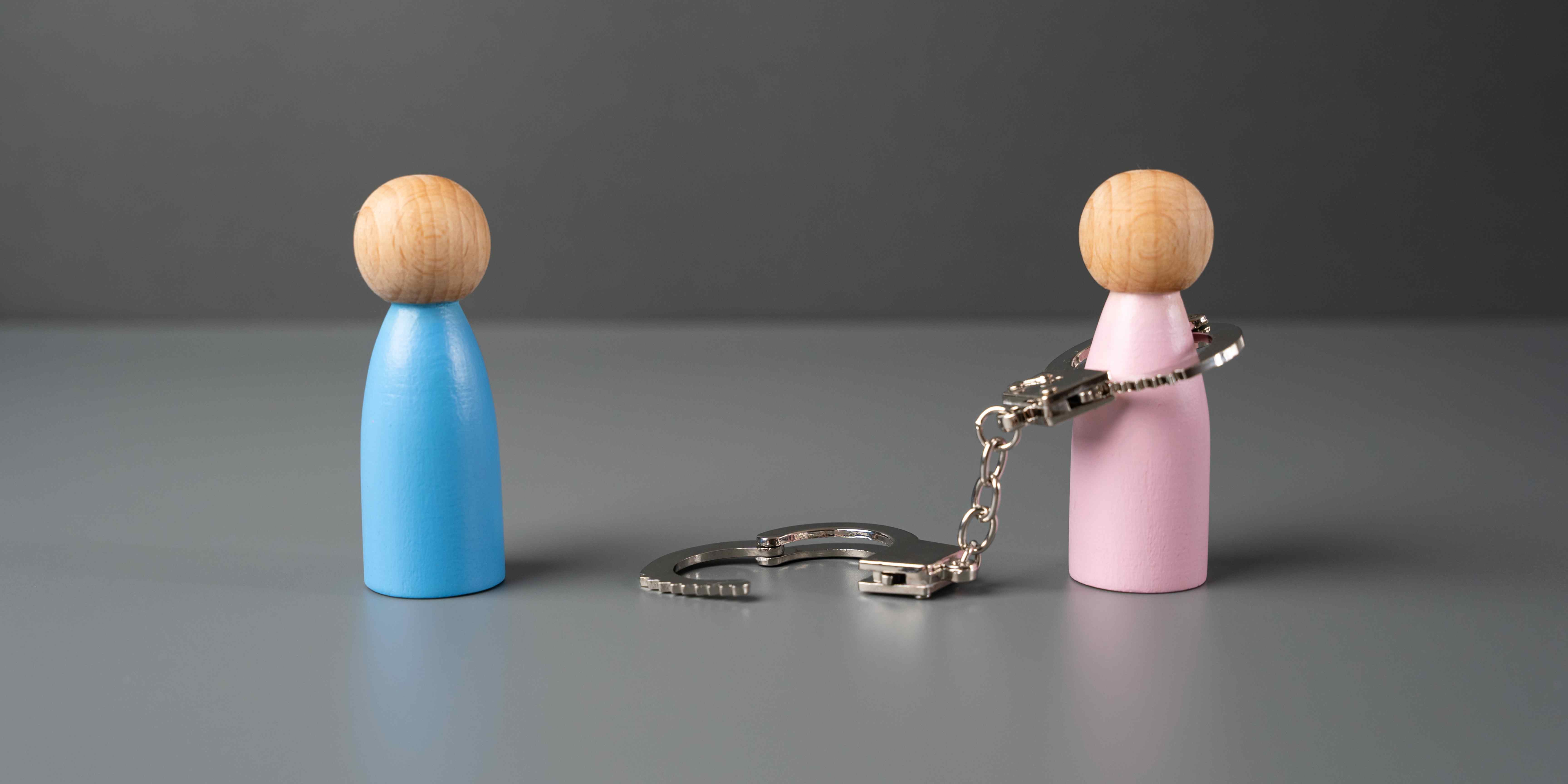 Two wooden figurines, one blue and one pink, stand apart. The pink figurine is attached to an open set of metal handcuffs—symbolizing the journey of healing from a toxic relationship—as the other cuff lies on the ground between them.