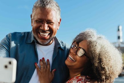 Smiling older couple taking a selfie together outdoors near the coast, with blue sky and a white lighthouse visible in the background. The woman wears glasses and the man is holding the phone.