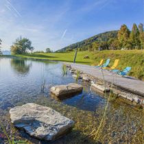 Experience tranquility at the Slovenia Yoga Retreat: Hiking, Swimming & more. Relax by a clear pond with rocks and tall grass; colorful lounge chairs line a wooden dock, surrounded by fields, trees, and scenic hills under a blue sky.