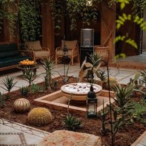A cozy courtyard features cactus plants, succulents, a decorative fountain with petals, lanterns, cushioned seating, and oranges—all set in a warm atmosphere inspired by the Morocco Yoga Retreat: Explore two magical.