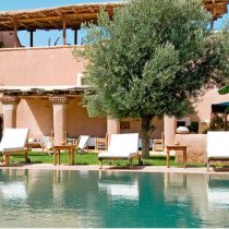Experience tranquility at the Morocco Yoga Retreat: Explore two magical destinations, where a serene outdoor pool with clear water is surrounded by white cushioned lounge chairs, lush greenery, and rustic architecture with a shaded terrace.