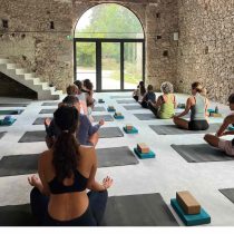 Participants of the Southwest France Yoga Retreat: An Exploration of Creativity and Rest practice yoga indoors on mats with blocks beside them, sitting cross-legged in a sunlit, rustic stone-walled room.