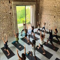 Participants in the Southwest France Yoga Retreat practice yoga in a rustic studio with stone walls and large windows, standing on mats and raising their arms in warrior pose while overlooking lush greenery.