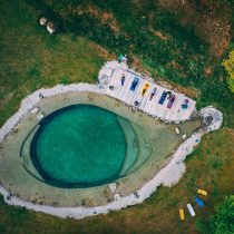 Aerial view of a human eye-shaped pond with a wooden deck where people practice yoga on mats, surrounded by green grass, trees, rocks, and colorful boats at the Slovenia Yoga Retreat: Hiking, Swimming & more.