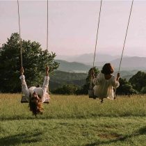 Two people in light outfits swing on wooden swings in a grassy field with mountains and trees behind them, evoking the natural setting of the Slovenia Yoga Retreat: Hiking, Swimming & more.