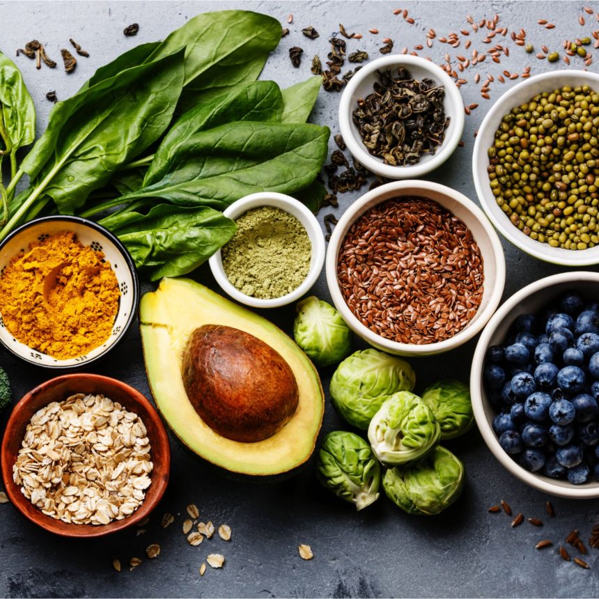 A variety of healthy foods including spinach, avocado, blueberries, Brussels sprouts, oats, flax seeds, mung beans, turmeric powder, green powder, and dried tea leaves, arranged on a gray surface.
