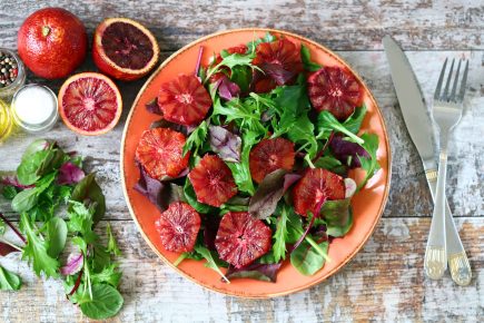 A plate of mixed green salad topped with blood orange slices sits on a rustic wooden table—perfect inspiration for anyone taking a free online nutrition class to learn about fresh, healthy eating. Nearby are whole and halved blood oranges, oil, salt, pepper, and utensils.