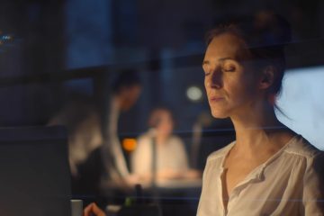 A woman sits at a desk with her eyes closed, appearing relaxed or meditative, in a dimly lit office. A laptop, plant, and office supplies are on the desk. Two blurred people work in the background.