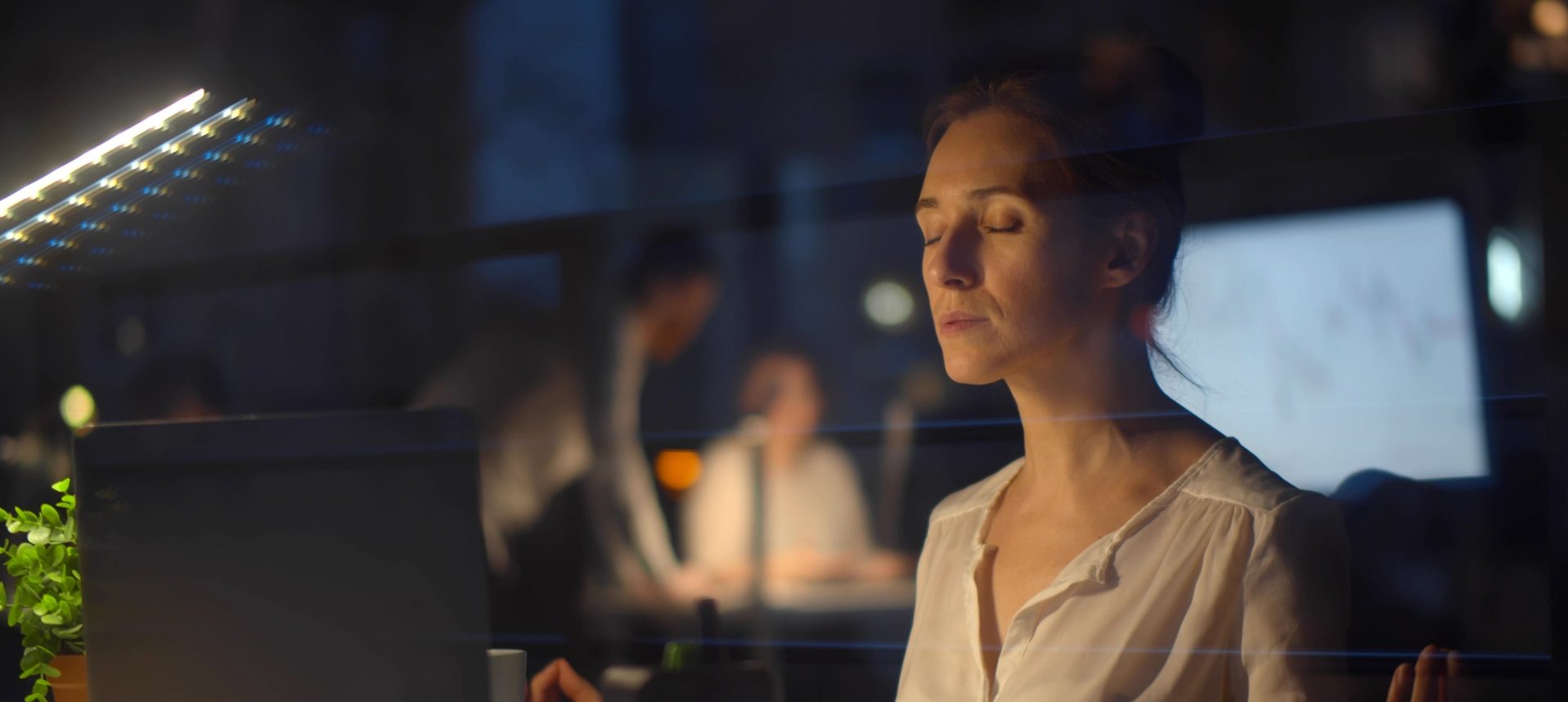 A woman sits at a desk with her eyes closed, appearing relaxed or meditative, in a dimly lit office. A laptop, plant, and office supplies are on the desk. Two blurred people work in the background.