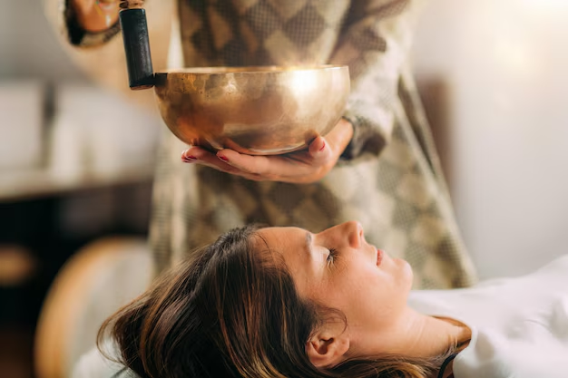 A person holds a singing bowl above a woman lying down with her eyes closed, suggesting a sound healing or relaxation session. Soft, warm light creates a calm and peaceful atmosphere.
