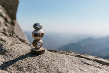 Three stones stacked on a rocky surface, each engraved with compass symbols, arrows, and the word Journey, with a blurred mountainous landscape in the background.