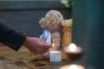 A person lights a candle near a teddy bear and a wicker basket, surrounded by more lit candles, creating a somber and reflective atmosphere.