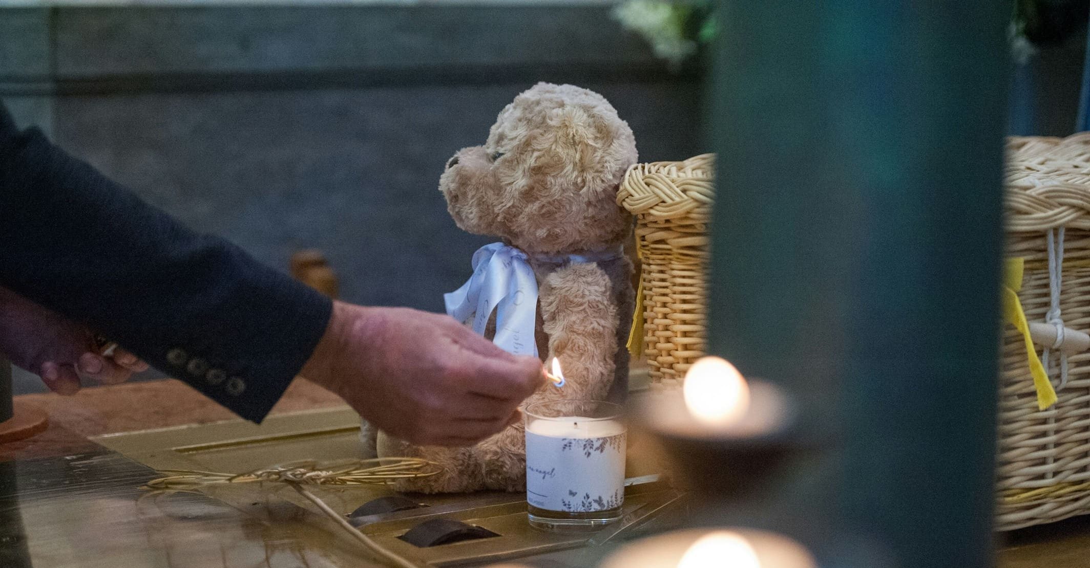 A person lights a candle near a teddy bear and a wicker basket, surrounded by more lit candles, creating a somber and reflective atmosphere.