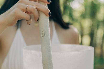 A woman in a white top holds a mallet above a frosted crystal singing bowl outdoors, surrounded by greenery.