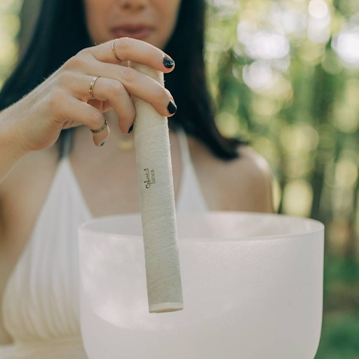 A woman in a white top holds a mallet above a frosted crystal singing bowl outdoors, surrounded by greenery.