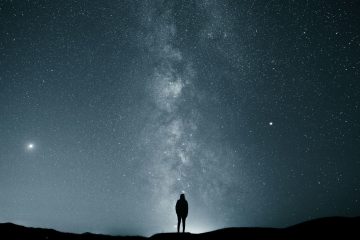 A person stands silhouetted on a hilltop at night, gazing up at a vast, star-filled sky with the Milky Way galaxy clearly visible above.