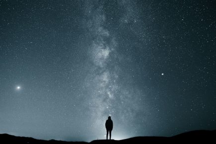 A person stands silhouetted on a hilltop at night, gazing up at a vast, star-filled sky with the Milky Way galaxy clearly visible above.