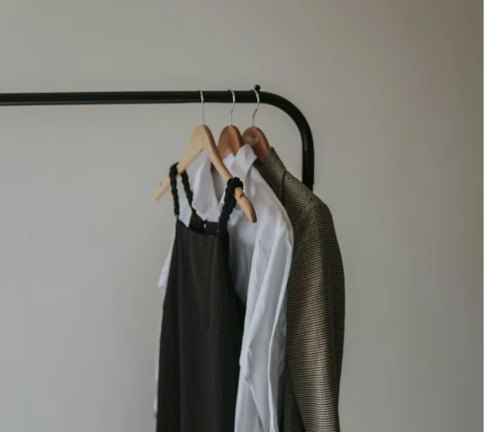 Three garments hang on wooden hangers from a black metal rack: a black dress, a white button-up shirt, and a patterned jacket. Their simple arrangement brings a sense of wellbeing to the space against the plain, neutral wall.