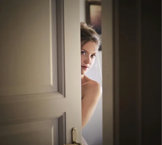 A woman with blonde hair partially peeks through a slightly open door, looking directly at the camera. Soft lighting creates a calm and intimate atmosphere, reflecting a sense of wellbeing in this plentiful, serene moment.