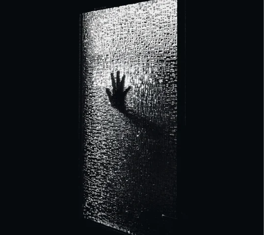 A silhouetted hand is pressed against a textured, frosted glass door, casting a shadow and creating a mysterious or eerie atmosphere—contrasting with the sense of wellbeing found in more plentiful light.