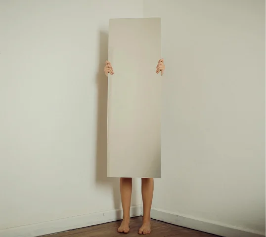 A person stands barefoot in a corner, holding a tall rectangular mirror that covers their body from shoulders to ankles, reflecting the blank wall behind them so their torso is hidden—suggesting a quiet search for wellbeing.