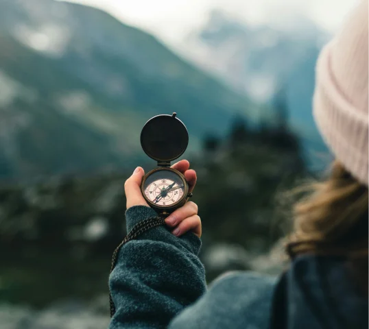 A person in a beanie and jacket holds an open compass, facing a blurred mountain landscape, suggesting outdoor exploration and the pursuit of plentiful wellbeing.