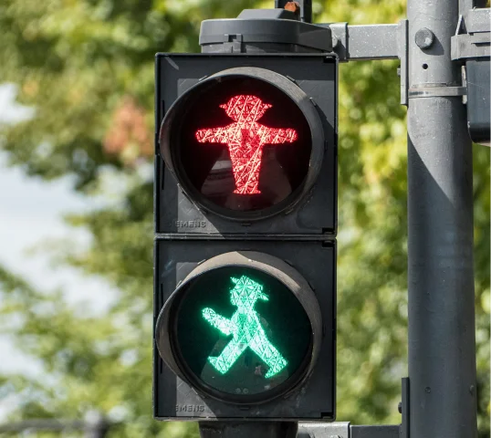A traffic light displays both a red standing figure and a green walking figure at once, giving conflicting signals for pedestrians. Plentiful trees and sky are visible in the background, adding a touch of natural wellbeing to the scene.
