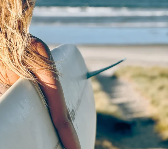 A person with long blonde hair holds a white surfboard while standing near the beach, embracing the plentiful sense of wellbeing that comes from ocean waves and sandy dunes in the background.