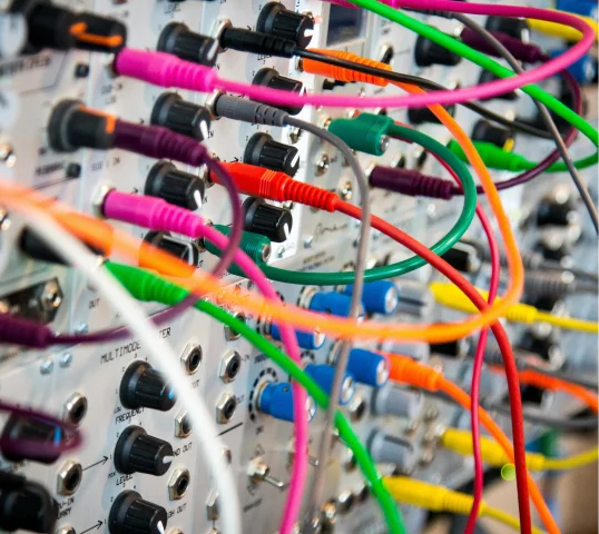 Close-up of a modular synthesizer with plentiful colorful cables plugged into various jacks and knobs, creating a complex network of connections on the control panel.
