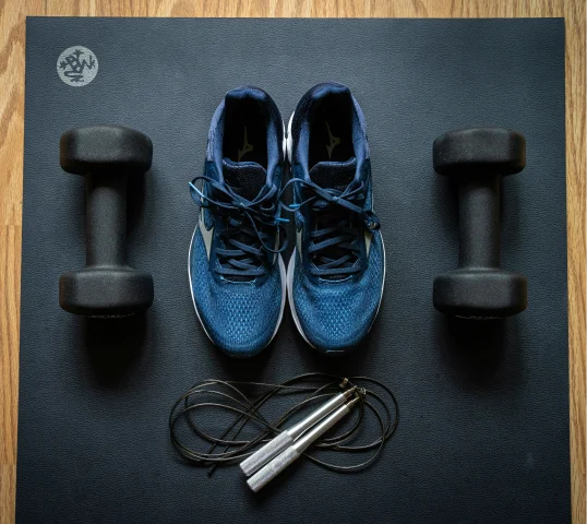 A pair of blue sneakers, two black dumbbells, and a black jump rope with silver handles are neatly arranged on a black exercise mat, creating a plentiful setup for your wellbeing on a wooden floor.