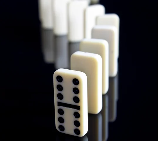 A row of white domino tiles is lined up vertically on a reflective black surface, with the front tile showing six dots above and below the center line, about to tip and start a plentiful chain reaction for wellbeing.