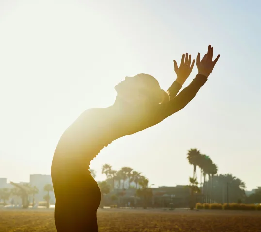 A person in athletic wear stretches backward with arms raised, silhouetted against a bright sun. Palm trees and buildings in the background evoke plentiful wellbeing in an outdoor, possibly beachside, location.