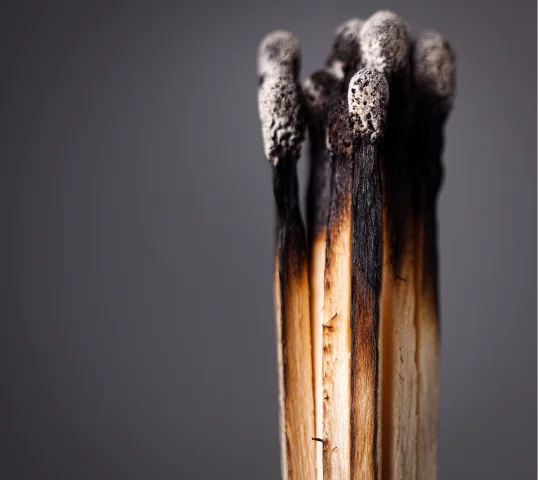 A close-up of several burnt matchsticks clustered together, their blackened, ashy tips and charred wood contrasting against a gray background—a striking reminder to protect your wellbeing before burnout replaces plentiful energy.