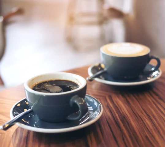 Two cups of coffee on saucers with spoons sit on a wooden table, embodying moments of plentiful wellbeing. One cup holds black coffee; the other, a creamy latte. The softly blurred background adds to this inviting sense of calm.