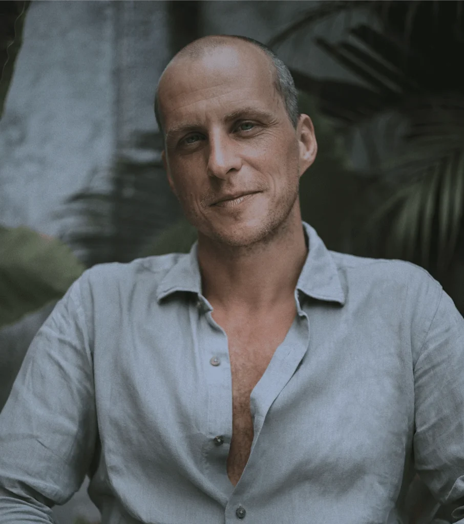 A middle-aged man with a shaved head and light stubble sits outdoors, wearing a light gray, partially unbuttoned shirt. He smiles gently, exuding wellbeing amidst greenery and soft sunlight in the blurred background.
