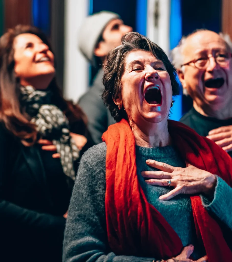 A group of older adults standing together indoors, hands on their chests, mouths open wide as if singing or laughing joyfully, wearing winter clothing and scarves—radiating plentiful wellbeing.