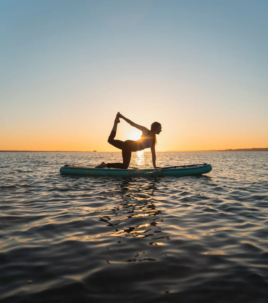 A person practices yoga on a paddleboard in calm water at sunset, balancing on one knee and holding one foot behind them—embracing wellbeing as the sun sets low on the horizon in the background.