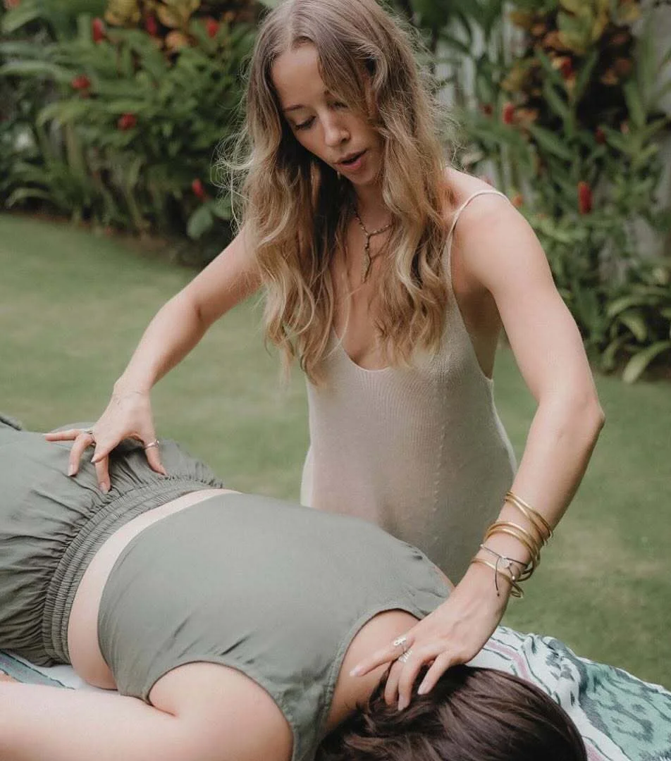 A woman with long wavy hair gives a therapeutic massage to another woman lying face down on a massage table outdoors, surrounded by plentiful lush green plants, creating an atmosphere of natural wellbeing.