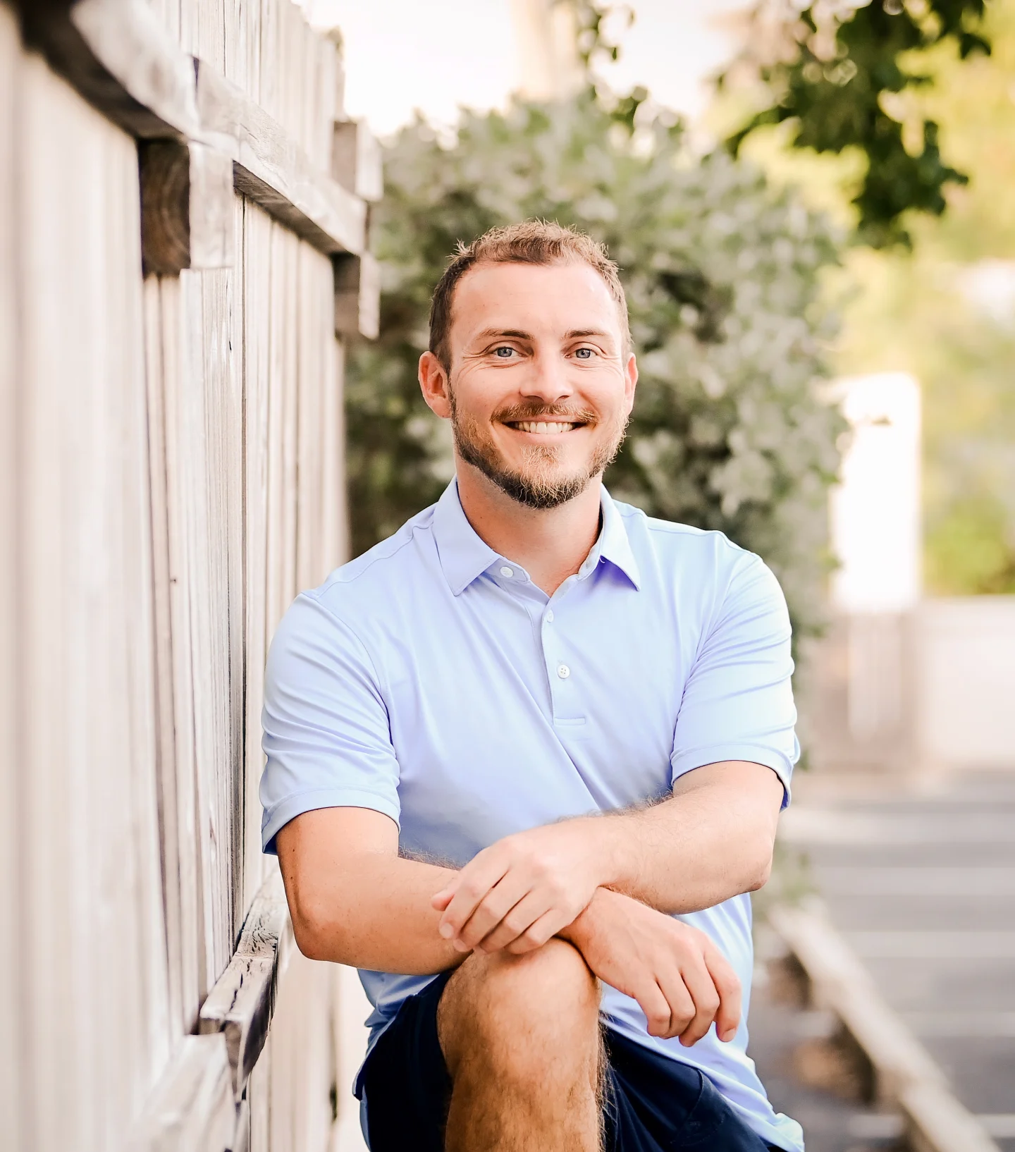 A smiling man with short hair and a beard, wearing a light blue polo shirt and dark shorts, sits outdoors by a wooden fence with greenery in the background on a sunny day, enjoying plentiful wellbeing.