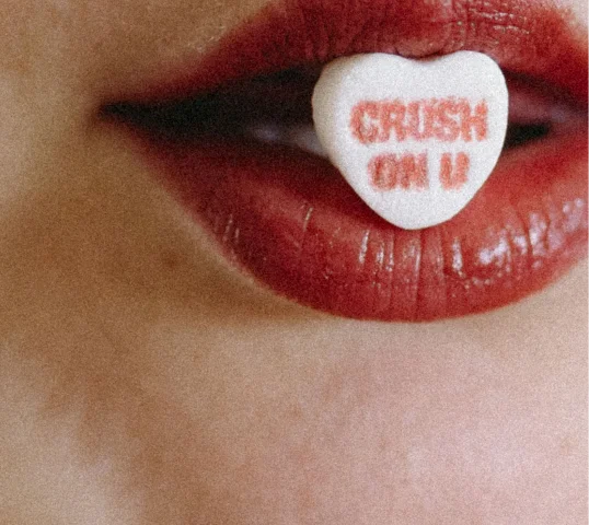 Close-up of red lips holding a heart-shaped candy between the teeth. The candy has red text that reads CRUSH ON U—radiating plentiful charm and sweetness, perfect for a boost to your online wellbeing.