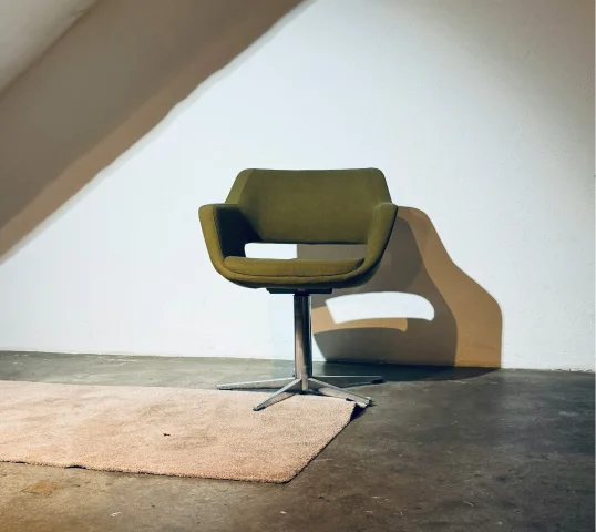 A modern olive-green swivel chair with metal legs sits on a bare concrete floor next to a beige rug, creating a tranquil space that promotes wellbeing and casts a shadow on a plain white wall.