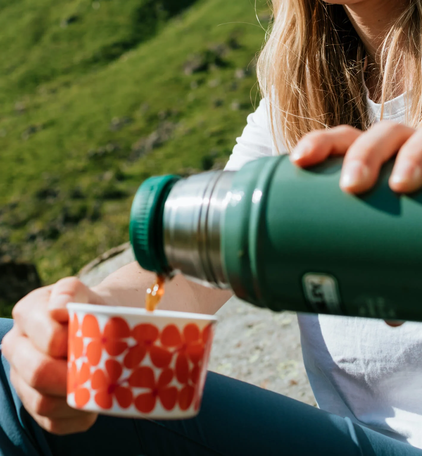 A person outdoors pours a drink from a green thermos into a red and white patterned cup, with green grass and sunlight in the background.