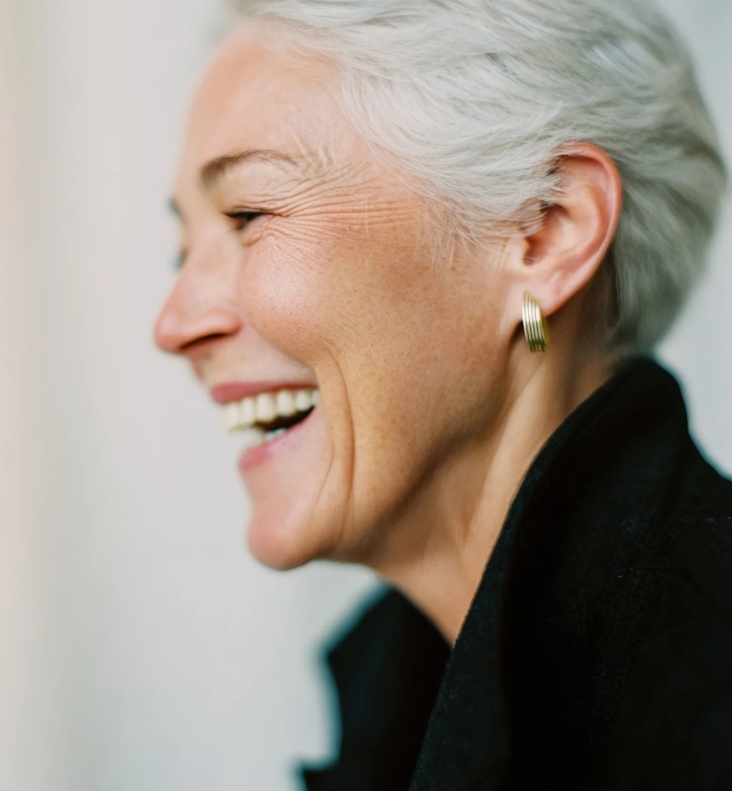 A joyful older woman with short white hair smiles and laughs, wearing a black jacket and a gold hoop earring, photographed in profile against a softly blurred background.