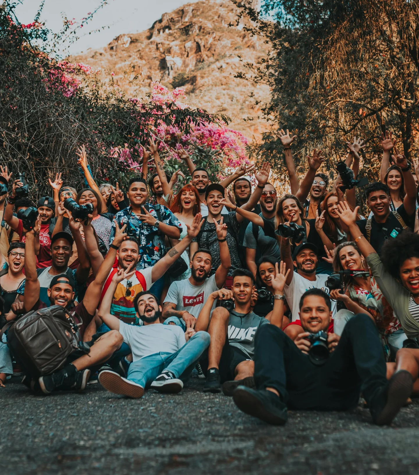 A large group of young adults sit and kneel outdoors on a path, smiling and raising their hands in excitement. Behind them are trees, pink flowers, and a rocky hillside under bright sunlight.